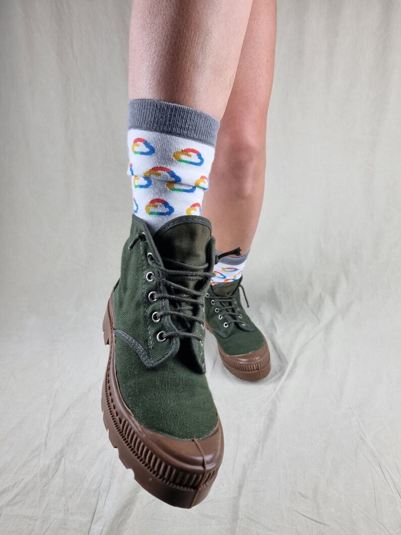 May include: Green canvas high-top boots with brown rubber soles and laces. The boots are worn with white socks featuring a colorful cloud pattern.