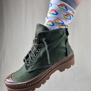 May include: Green canvas high-top boots with brown rubber soles and a thick tread. The boots are worn with white socks featuring a colorful pattern.