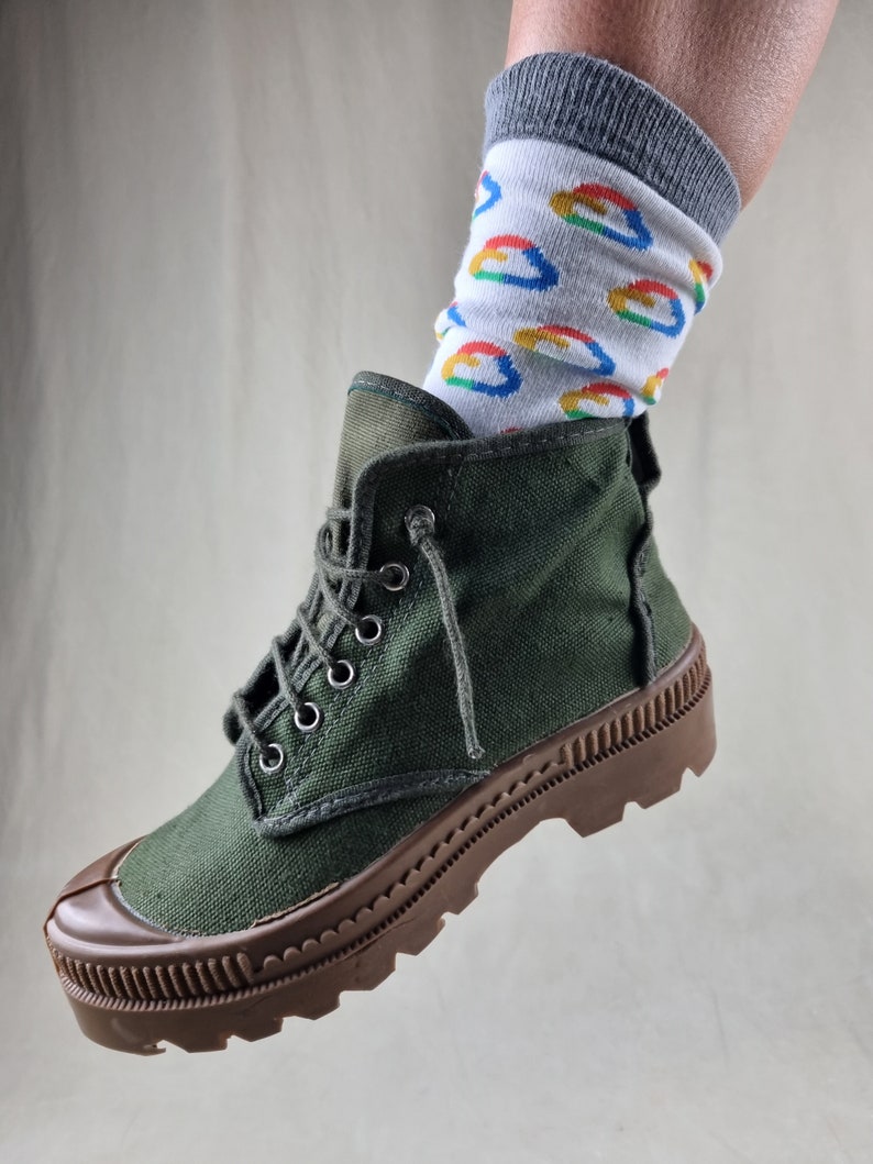 May include: Green canvas high-top boots with brown rubber soles and a thick tread. The boots are worn with white socks featuring a colorful pattern.