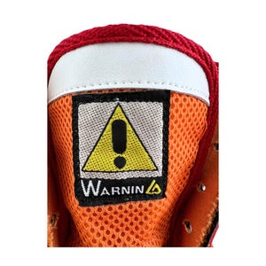 May include: Close-up of a red and orange shoe with a yellow warning sign that says "Warning" with an exclamation point inside a triangle.