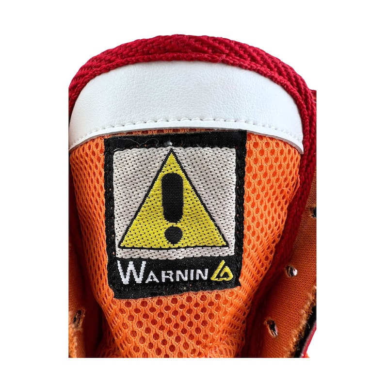 May include: Close-up of a red and orange shoe with a yellow warning sign that says "Warning" with an exclamation point inside a triangle.