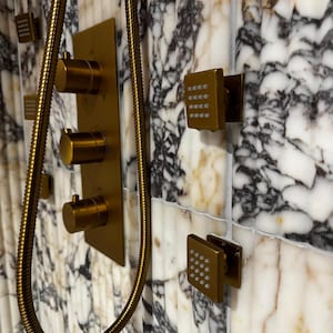 May include: A gold-toned shower fixture set against a marble tile wall. The set includes a control panel with three knobs, a flexible shower hose, and multiple square shower heads with small spray holes.
