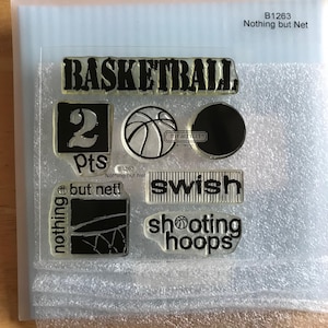 May include: A set of clear stamps with black ink featuring basketball-themed images and text, including "BASKETBALL", "2 pts", "but net!", "swish", and "shooting hoops".