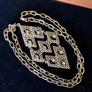 May include: A gold chain necklace with a large, geometric pendant made of interlocking squares. The pendant has a delicate, filigree design.