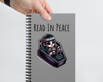 Może przedstawiać: Szary notes w spirali z tekstem "READ IN PEACE" i kreskówkową ilustracją dziewczyny w trumnie czytającej książkę.