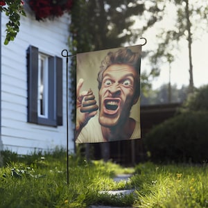 May include: A garden flag featuring a digitally altered image of a man with a shocked expression, holding up a pile of feces. The flag is displayed outdoors in a grassy yard.