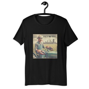 May include: Black t-shirt with a graphic of a person with a lobster head driving a tractor in a field with a city skyline in the background. The text "ONLY IN OHIO" is above the city skyline.