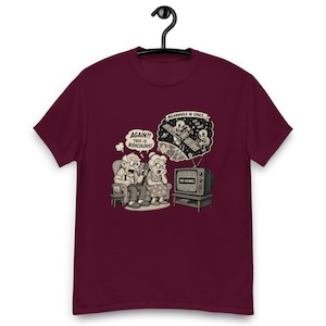 Könnte beinhalten: Burgunderfarbenes T-Shirt mit einem humorvollen Cartoon-Design. Die Grafik zeigt ein älteres Ehepaar, das auf einen "No Signal"-Fernsehbildschirm reagiert, mit einer Weltraumszene darüber. Text enthält "AGAIN?! THIS IS RIDICULOUS!" und "MEANWHILE IN SPACE."