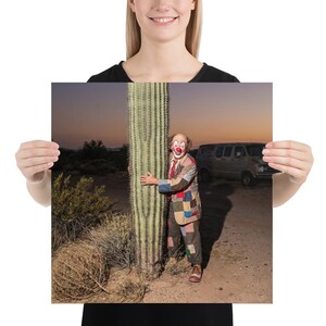 May include: A print featuring a clown embracing a tall cactus in a desert landscape. The clown is wearing a colorful patchwork suit, red tie, and clown makeup. A vintage van is in the background, and the sky is a gradient of orange and purple.