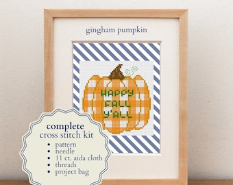 May include: A complete cross stitch kit featuring a gingham pumpkin design with the text "HAPPY FALL Y'ALL" in green. The kit includes a pattern, needle, 11 ct. aida cloth, threads, and a project bag, all displayed in a light wood frame.
