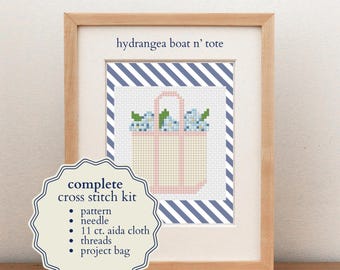 May include: A framed cross stitch kit titled hydrangea boat n' tote featuring a beige tote bag with pink handles and blue hydrangea flowers. The kit includes a pattern, needle, 11 ct. aida cloth, threads, and a project bag, all displayed in a light wood frame.