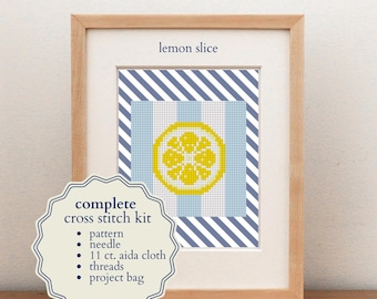 May include: A complete cross stitch kit featuring a lemon slice design in a light wood frame. The kit includes a pattern, needle, 11 ct. aida cloth, threads, and a project bag. The cross stitch displays a yellow lemon slice on a light blue and white striped background with a dark blue and white diagonal striped border.
