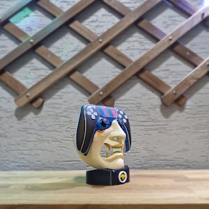 May include: A unique sculpture featuring a face with a controller design. The face is cream-colored with a detailed open mouth, and the controller has a black, blue, and colorful design. It sits on a black base with a gold emblem.