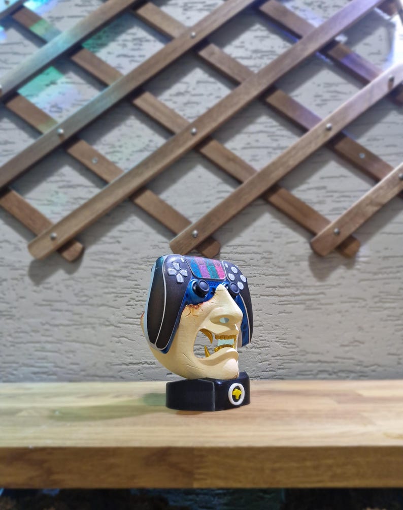 May include: A unique sculpture featuring a face with a controller design. The face is cream-colored with a detailed open mouth, and the controller has a black, blue, and colorful design. It sits on a black base with a gold emblem.