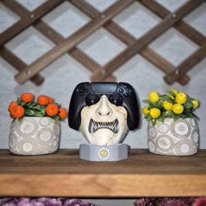 May include: A black video game controller rests on a white and gray decorative stand shaped like a Japanese demon mask. Two small potted plants with orange and yellow fruit flank the controller on a wooden shelf.