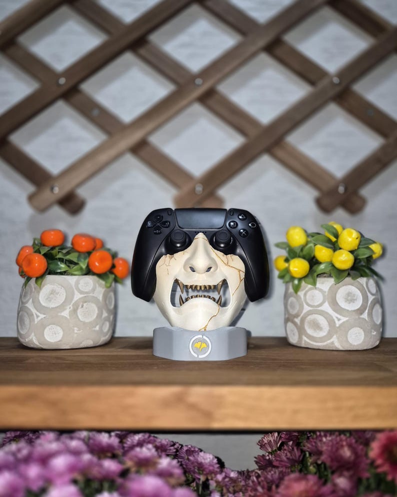 May include: A black video game controller rests on a white and gray decorative stand shaped like a Japanese demon mask. Two small potted plants with orange and yellow fruit flank the controller on a wooden shelf.