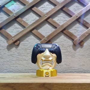 May include: A unique gaming controller holder featuring a stylized face design with a black controller integrated into the top. The holder is cream-colored with gold accents and sits on a gold base.