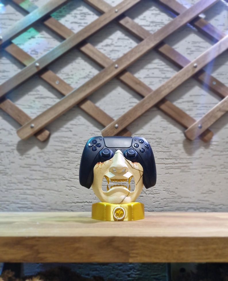 May include: A unique gaming controller holder featuring a stylized face design with a black controller integrated into the top. The holder is cream-colored with gold accents and sits on a gold base.