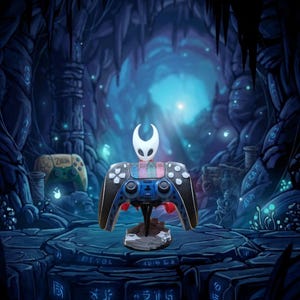 May include: A black, blue, and red video game controller on a stand, with a white character figure on top. The controller is set against a blue and black cave background, with a Zelda controller in the background.