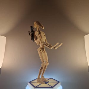 May include: A beige and white Star Wars action figure of a battle droid standing on a hexagonal base with a blue light. The droid is holding a blaster rifle in its right hand.