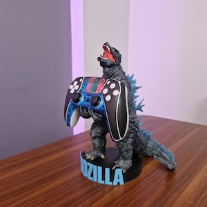 May include: A blue and black video game controller is held by a Godzilla figurine. The figurine is standing on a black base with the word "Godzilla" printed in blue letters.