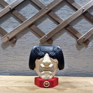 May include: A unique decorative item featuring a black game controller atop a stylized face sculpture. The face is off-white with a cracked design, set on a red base with a circular emblem. The background includes a wooden trellis.