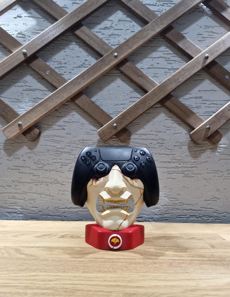 May include: A unique decorative item featuring a black game controller atop a stylized face sculpture. The face is off-white with a cracked design, set on a red base with a circular emblem. The background includes a wooden trellis.
