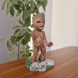 May include: A brown figurine of Groot from Guardians of the Galaxy, with an angry expression and open mouth. The figure stands on a green base with gray rock accents. The figure is approximately 5 inches tall.