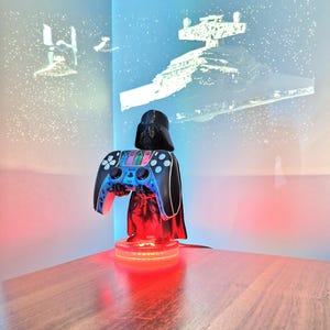 May include: Darth Vader figurine holding a black and blue video game controller. The figurine is on a circular base with red LED lights. The background is a blue and white wall with a projection of a Star Wars spaceship.