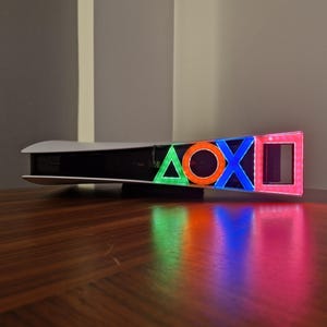 May include: A white gaming console with a black stripe and four colored lights in the shape of a triangle, circle, cross, and square. The lights are green, orange, blue, and red.