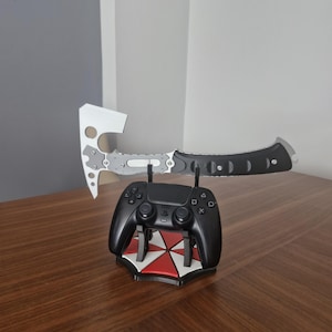 Pode incluir: Um comando de videojogos preto repousa sobre um suporte com um logótipo de guarda-chuva vermelho e branco. Um machado prateado e preto com um design recortado está equilibrado horizontalmente sobre o comando, exibindo uma lâmina e cabo metálicos.