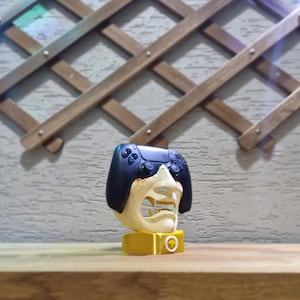 May include: A unique, yellow and off-white gaming controller stand shaped like a stylized face with a black controller on top. The stand is on a wooden surface with a decorative wooden trellis in the background.
