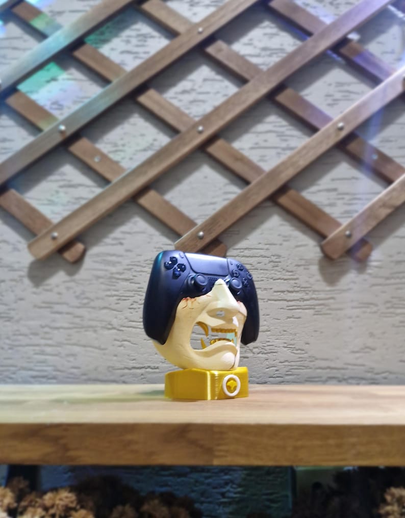 May include: A unique, yellow and off-white gaming controller stand shaped like a stylized face with a black controller on top. The stand is on a wooden surface with a decorative wooden trellis in the background.