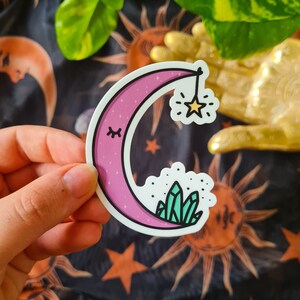 Könnte beinhalten: Ein Aufkleber mit einem rosa Halbmond mit geschlossenem Auge, weißem Umriss und einem hängenden Stern. Das Monddesign beinhaltet eine Wolke mit grünen Kristallen. Der Aufkleber wird von einer Hand gehalten.
