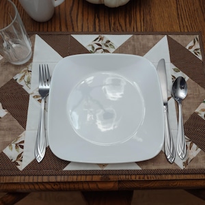 May include: A place setting on a wooden table, featuring a white square plate, silverware, and a patterned placemat. The placemat has a brown and cream geometric design with leaf accents. A clear glass and a white mug are also visible.