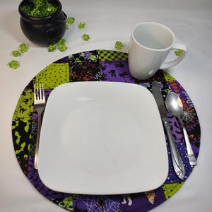 May include: A purple, green, and black Halloween-themed placemat with bats, spiders, and the words "Happy Halloween" and "RIP". A white square plate, a white mug, a fork, a knife, and a spoon are on top of the placemat.