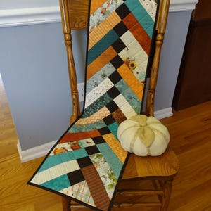 May include: A colorful, quilted table runner with a black border is draped over a wooden chair. The runner features a patchwork design with teal, orange, brown, and black squares. A white pumpkin with a tan stem sits on the chair.
