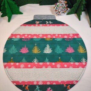 May include: A green and pink Christmas ornament-shaped placemat with a white snowflake pattern and embroidered Christmas trees. The placemat is made of fabric and has a stitched edge.