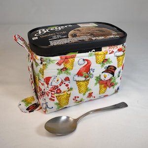 May include: A white fabric ice cream container holder with a snowman and Santa Claus design. The holder is filled with a Breyers Chocolate Truffle ice cream container and a silver spoon is on the white surface.