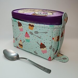 May include: A quilted fabric ice cream pint cozy in light blue with a pattern of cupcakes, ice cream scoops, and spoons, featuring a side tab closure. The cozy holds a purple plastic container labeled PURPLE PREMIUM ICE CREAM. A metal spoon lies nearby.