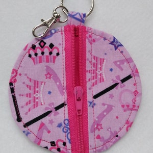 May include: A round, pink fabric keychain wallet with a zipper closure. The wallet features a pattern of crowns, stars, and magic wands in shades of pink and purple. It has a silver key ring and a lobster clasp for easy attachment.
