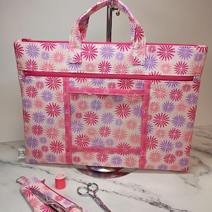 May include: A rectangular fabric bag featuring a repeating pattern of pink and purple daisy-like flowers on a cream background, with a top zipper, two handles, and a front pocket. A matching small fabric case, scissors, and pink thread are included.