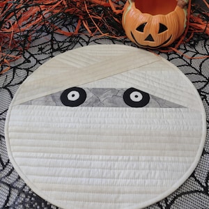 May include: A round, quilted placemat with a mummy design for Halloween. The placemat is cream-colored with horizontal stitching and a gray band with black eyes. A ceramic jack-o'-lantern basket sits nearby.