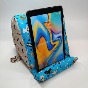May include: A blue tablet pillow stand with a dog and bone pattern, holding a black Samsung Galaxy Tab A tablet. The pillow has a light tan side with a bone pattern. The tablet displays a colorful abstract image.