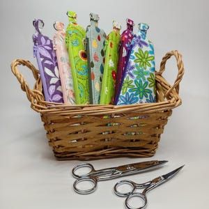 May include: A collection of seven rectangular fabric zipper pouches with various colorful floral and patterned prints, each featuring a metal D-ring at the top, displayed in a woven wicker basket with two silver metal scissors placed in front.