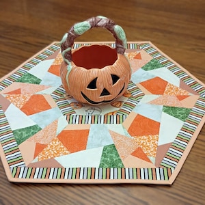 May include: A ceramic jack-o'-lantern basket with a vine-like handle sits atop a hexagonal fabric mat. The mat features a patchwork design of orange, green, and white geometric shapes with a striped border in black, white, orange, and green.