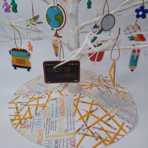 May include: A white artificial tree decorated with school-themed ornaments, including a school bus, a globe, a magnifying glass, a pencil, a ruler, an apple, and a chalkboard with the text "Back to School". The tree is sitting on a white base with a yellow pencil pattern and school-related words printed on it.