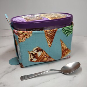 May include: An ice cream cozy with a light blue background and ice cream cone pattern. The cozy holds a purple ice cream container. A silver spoon rests on the surface next to the cozy.