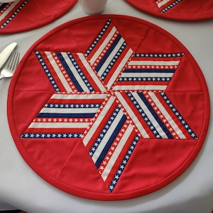 May include: Red, white, and blue fabric placemat with a star design. The placemat is round and has a red border.
