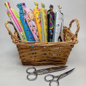 May include: A woven wicker basket containing seven tall, rectangular fabric pouches with zippers and metal rings at the top, featuring various colorful patterns including sewing themes. Two silver metal scissors lie in front of the basket.